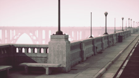 Morning fog envelops a serene promenade. Old lampposts line the pathway, while benches invite quiet reflection. In the background, a grand bridge looms faded and mysterious in the mist.の写真素材