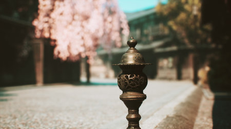 A detailed metal lantern features intricate carvings, placed on a serene pathway. Cherry blossoms surround the area, creating a peaceful and picturesque scene on a bright day.の写真素材