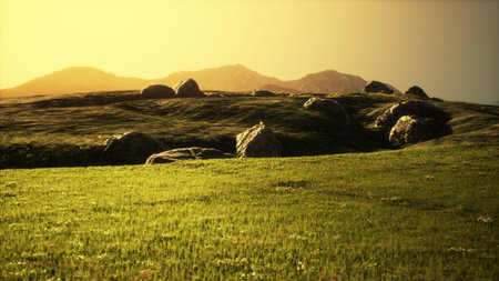 As the sun sets on the horizon, vibrant hues paint the sky while gentle hills covered in lush grass present a tranquil scene embellished with large, scattered boulders.の写真素材