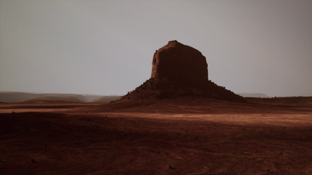 In the quiet twilight, a solitary rock formation rises from the arid desert, surrounded by golden sands and distant hills. Shadows stretch across the terrain as night approaches.の写真素材