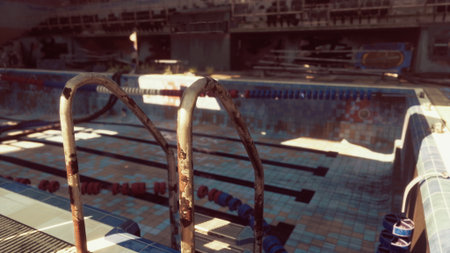 An old indoor swimming pool sits empty, sunlight filters through dusty windows highlighting the rusted metal ladder. Tiles are cracked, and lanes are marked with faded colors.の写真素材