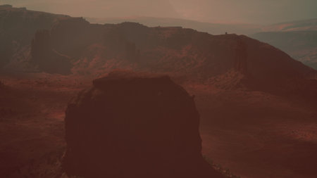 A breathtaking scene unfolds with vast red rock formations casting long shadows as the sun sets behind distant mountains. The hazy atmosphere adds mystery to the deserts rugged beauty.の写真素材
