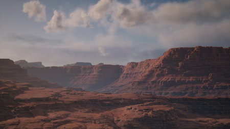 The sun sets over expansive red rock formations, casting warm hues on the rugged terrain. Clouds drift lazily above, adding beauty to the tranquil desert scenery.の写真素材