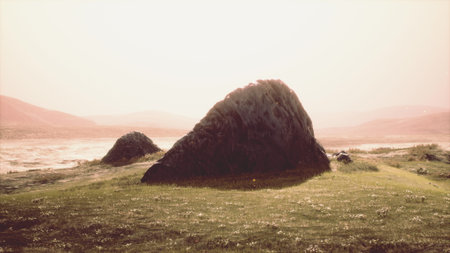 A large rock stands majestically on a green field, illuminated by soft sunlight. The tranquil scenery features distant hills and a calm water body, creating a peaceful atmosphere during the day.の写真素材