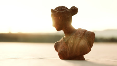 A clay bust of a woman captures sunlight as it sets in the background. The warm glow highlights the intricate details of the sculpture. Nature creates a calm and reflective atmosphere.の写真素材