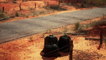 Bags of waste are left beside a barren road in a vibrant red landscape. The scene highlights the contrast between nature and human impact. A dry environment and minimal vegetation surround the area.の写真素材