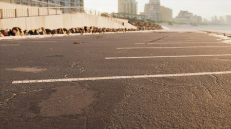 A cracked asphalt parking lot stretches along the beach, reflecting the warm hues of sunset. Buildings rise in the background, creating a calm urban coastal scene.の写真素材