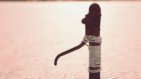 A vintage water pump stands by a calm lake as the sun sets, casting beautiful warm hues on the water...