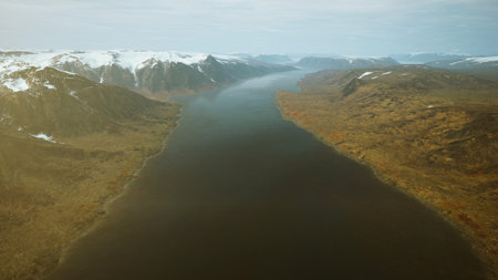 A stunning landscape showcases a wide river meandering through rugged terrain, flanked by snow capped mountains and golden autumn foliage. The atmosphere is serene and peaceful.の写真素材