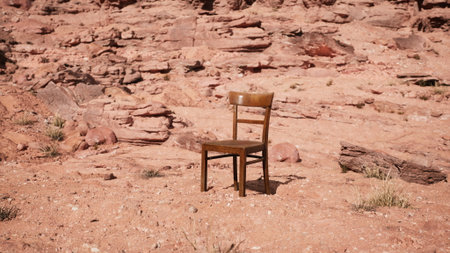 A wooden chair rests alone on the sandy ground of a rugged desert landscape, surrounded by colorful rocks and sparse vegetation, bathed in warm sunlight during the day.の写真素材