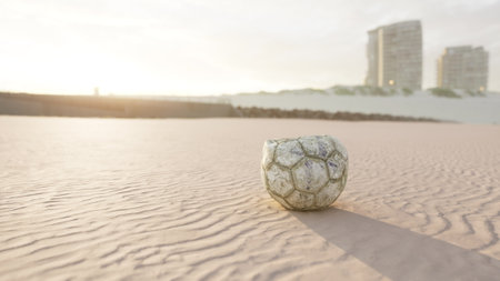 A worn soccer ball sits on a smooth sandy beach as the sun sets behind distant buildings. The light casts long shadows, creating a serene atmosphere on the shore.の写真素材