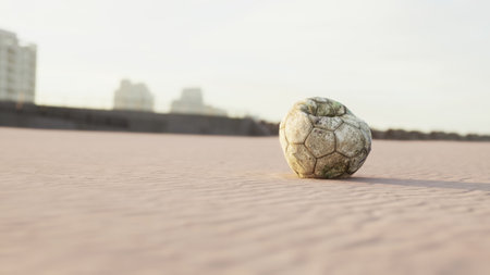A worn soccer ball rests on soft sand, surrounded by an empty beach. The distant city skyline glows softly in the evening light, creating a peaceful yet nostalgic scene.の写真素材