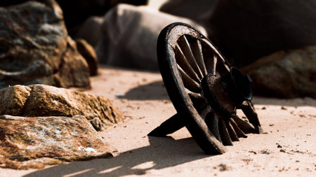 A weathered wooden wheel leans against soft sand, surrounded by rustic rocks. The sun sets, casting a warm glow and creating shadows that tell a story of time and natures beauty.の写真素材