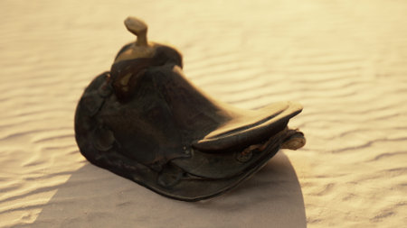 A weathered leather saddle lies abandoned on soft desert sand as the sun sets. The warm, golden light enhances the textures and history of this rustic equipment.の写真素材