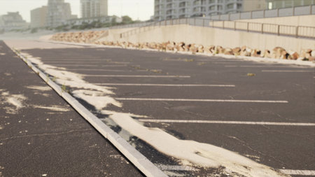 A coastal parking lot shows signs of erosion as sand spreads across the pavement. Waves have washed ashore, creating natural designs that blend land and sea.の写真素材
