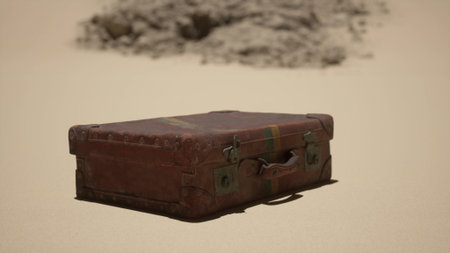 A vintage suitcase sits on golden sand, surrounded by vast desert. The sun illuminates its worn leather, showing signs of age. Nearby, a distant rocky formation adds to the desolate beauty.の写真素材