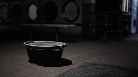 In a dimly lit laundromat, a solitary black bucket sits silently on the tile floor, surrounded by shadows that create an air of mystery and intrigue.の写真素材