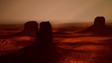 Towering buttes stand silently, bathed in warm orange light at sunset, casting long shadows across the vast desert terrain. The scene inspires tranquility and awe.の写真素材