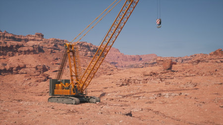 A towering crane stands in a vast desert with rocky formations and red sand. Its arm stretches high as it prepares to lift a heavy load against a backdrop of distant mountains.の写真素材