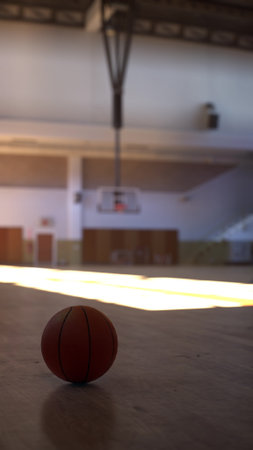 A basketball rests on the court with soft, warm light illuminating the gym. Shadows stretch across the wooden floor, creating a serene and inviting atmosphere during a quiet moment.の写真素材