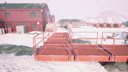 Pathways made of metal connect red buildings in a snow covered area. Nearby, mountains rise against a pale sky, highlighting the remote beauty of this chilly landscape.の写真素材