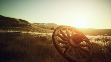 A weathered wagon wheel rests in a golden field as the sun sets on the horizon. The warm light casts a magical glow over the tranquil landscape, evoking a sense of nostalgia and peace.の写真素材