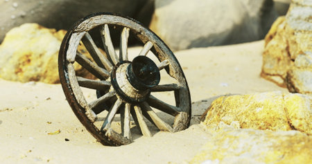 A wooden cart wheel rests partially buried in soft sand, surrounded by rocks. The setting suggests a deserted coastal area, highlighting the contrast between nature and manmade objects.の写真素材