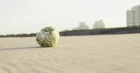 A weathered soccer ball sits on a sandy beach, partially buried in the grains. In the distance, tall buildings create a vibrant urban backdrop under a clear sky.の写真素材