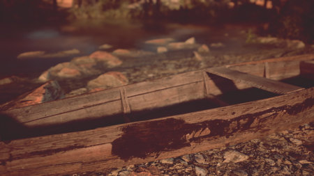 A weathered wooden boat lies on the shore of a peaceful lake as the sun sets, casting warm light on the surrounding rocks and water, creating a serene evening atmosphere.の写真素材