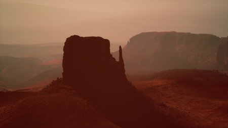 A majestic rock formation stands prominently against a sunset sky, casting deep shadows on the surrounding desert. The warm colors create a serene yet mysterious atmosphere.の写真素材