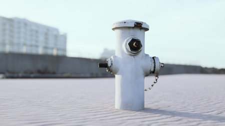 A white fire hydrant stands prominently on a sandy area, with a backdrop of distant city buildings....