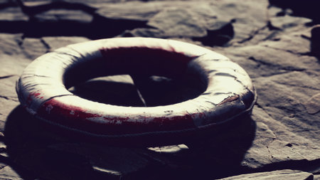 A weathered lifebuoy lies on rough rocks near the shore as the sun sets, casting warm hues across the scene. Waves gently lap at the edge, creating a serene coastal moment.の写真素材