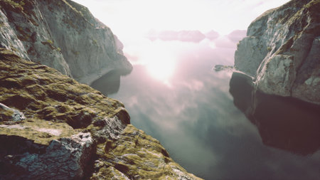 Calm waters mirror dramatic cliffs and distant peaks as the sun rises, casting a gentle glow on the landscape. Peaceful and untouched, natures beauty enchants the viewer.の写真素材