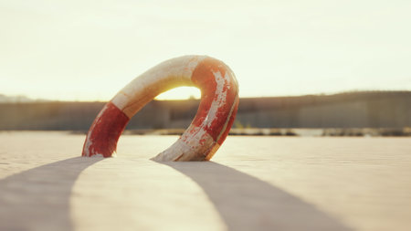 A vibrant lifebuoy lies on golden sand, casting a long shadow. The sun sets in the background, illuminating the tranquil atmosphere and reflecting on calm waters nearby, creating a serene scene.の写真素材