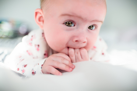 Four months old baby girl on a bed looking at camera in sunny bedroom. Fingers in mouth.