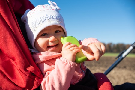 Adorable baby girl outside in red stroller in green fields on a road, sunny day. Baby with green toy...