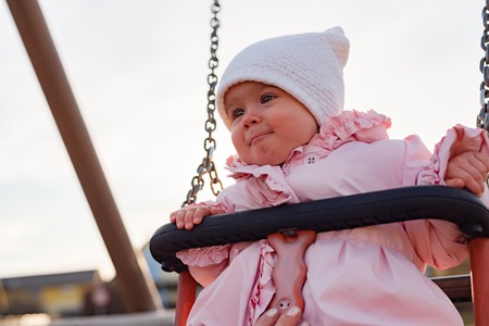Adorable baby girl with big beautiful eyes and a beanie having fun on a swing ride at a playground in a sunny parkの写真素材