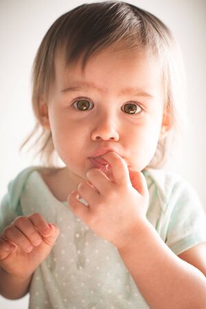 Cute caucasian 1 and half year old baby girl puts fingers into her mouth, concept of teething.