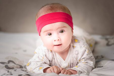Adorable 6 months old Baby girl infant on a bed on her belly with head up looking into camera with her big eyes. Natural bedroom light.の写真素材