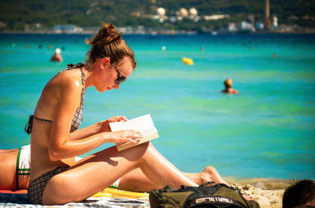 Alcudia, Spain 14.09.2011 - Young woman reading a book at sandy beach. People sunbathing at Playa de Muro. Mallorca island famous tourist destination.のeditorial素材
