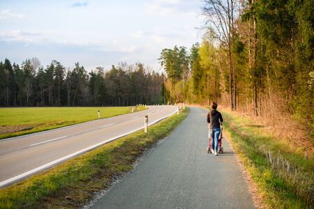 Young mother and child in the red stroller walks on path allong street in rural area before sunset. Familly time conceptの写真素材