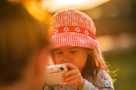 Portrait of a beautiful little 2 years old girl outdoors in red hat shooting photos with camera. Sunset light backgroundの写真素材