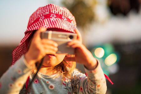 Portrait of a beautiful little 2 years old girl outdoors in red hat shooting photos with camera. Sunset light backgroundの写真素材