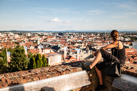 Graz, Austria - 13.07.2020: Woman sitting on red wall against panorama view from the top of schlossberg hill over the city. Selectiv focus on womanのeditorial素材