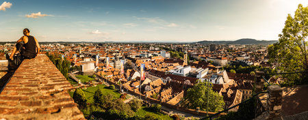 Graz, Austria - 13.07.2020: Woman sitting on red wall against panorama view from the top of schlossberg hill over the cityのeditorial素材