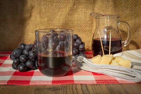 glass of red wine with jug, bunch of grapes and pieces of parmesan cheese on table with white and red checkered tableclothの写真素材