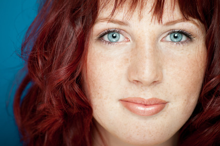 Beautiful fresh girl with auburn hair, blue eyes and freckles posing over blue background.の写真素材