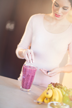 Young European woman making fresh berry smoothie in kitchen. Healthy eating concept. Full glass of healthy juice.の写真素材