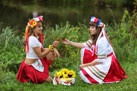 Two girls in Ukrainian costumes dressed for the holiday of Ivan Kupala sitting on the Bank of the river and drink vodkaの写真素材