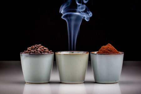 3 rustic coffee cups with coffee grounds, smoking coffee, and ground coffee. Standing on a white reflective table fading into a black backgroundの写真素材